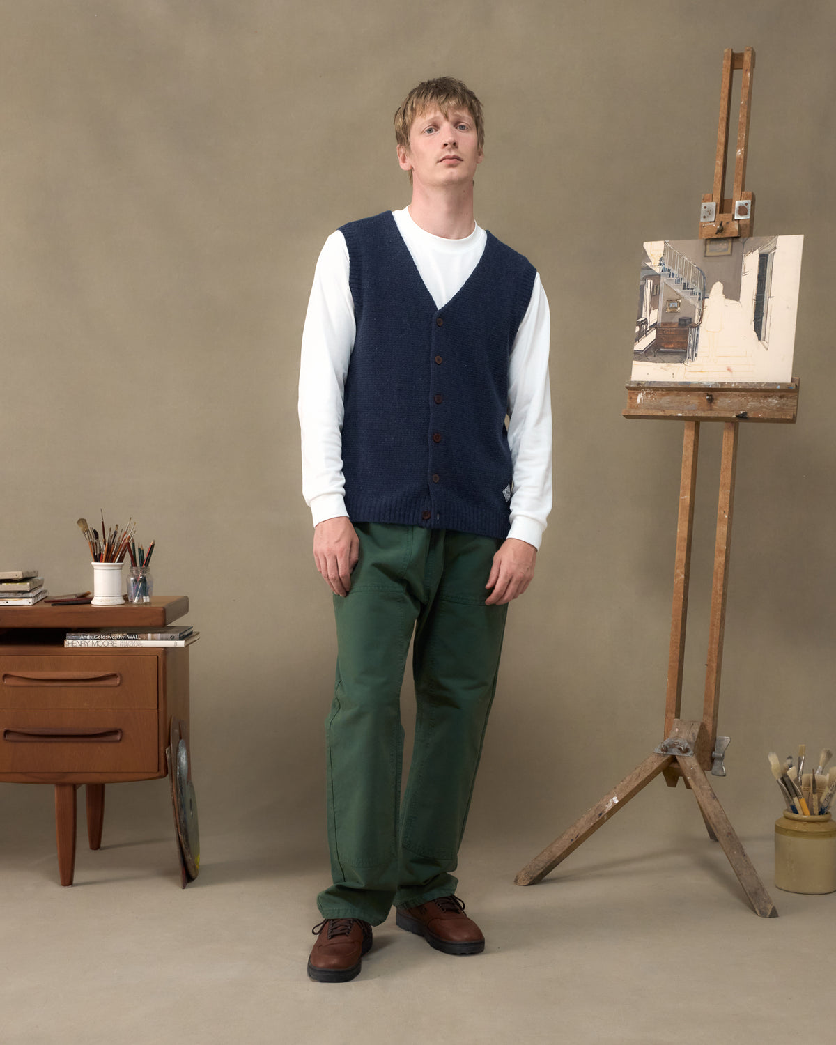 Model wearing the Fera Curlew Merino Vest in Navy with green trousers beside a painter's easel