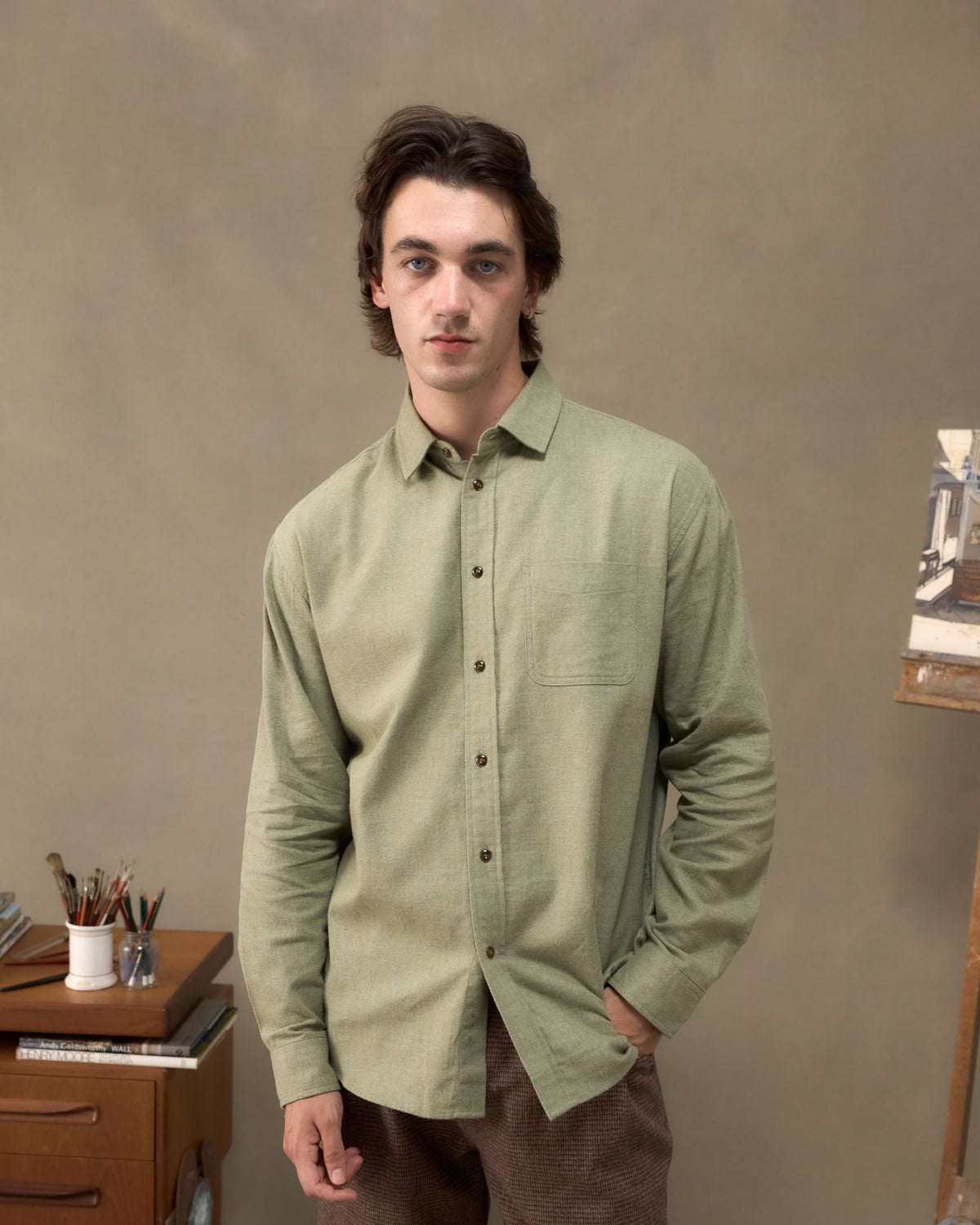 Model wearing the Fera Avon Shirt in Brushed Green, front view beside a painter's desk