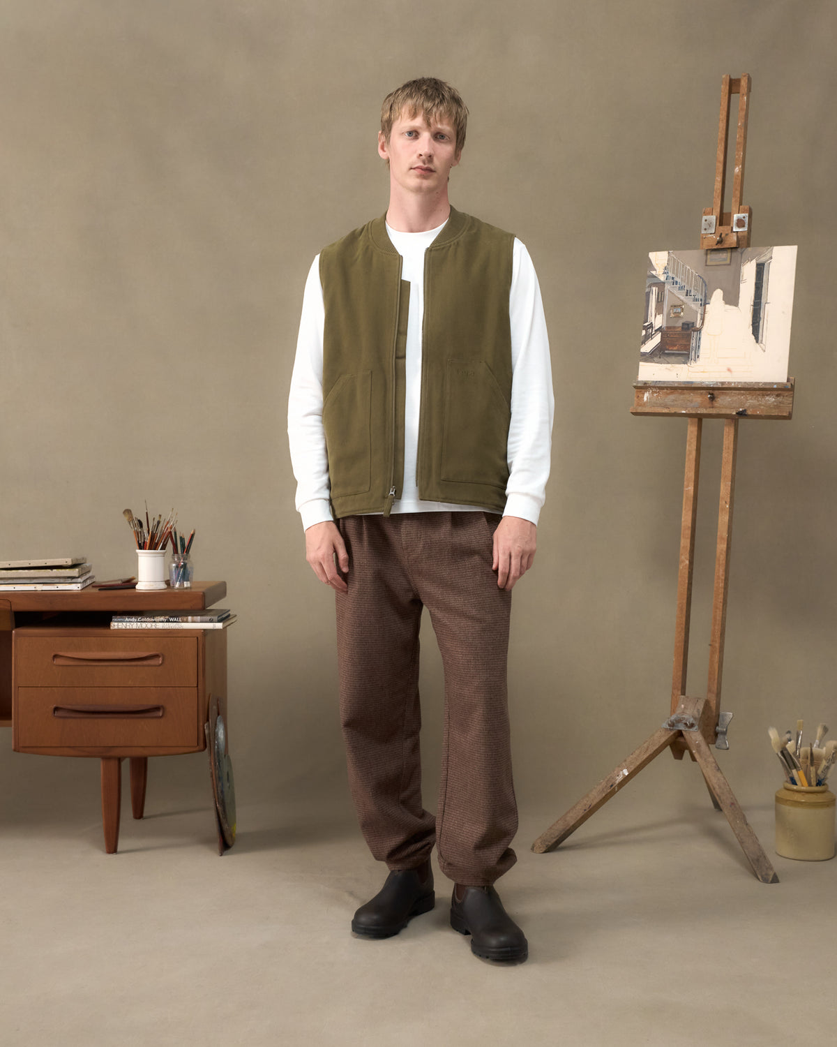 Model wearing the Fera Poacher Gilet in Marsh Green with Fera Uniform Trousers in Tweed in an artist's studio
