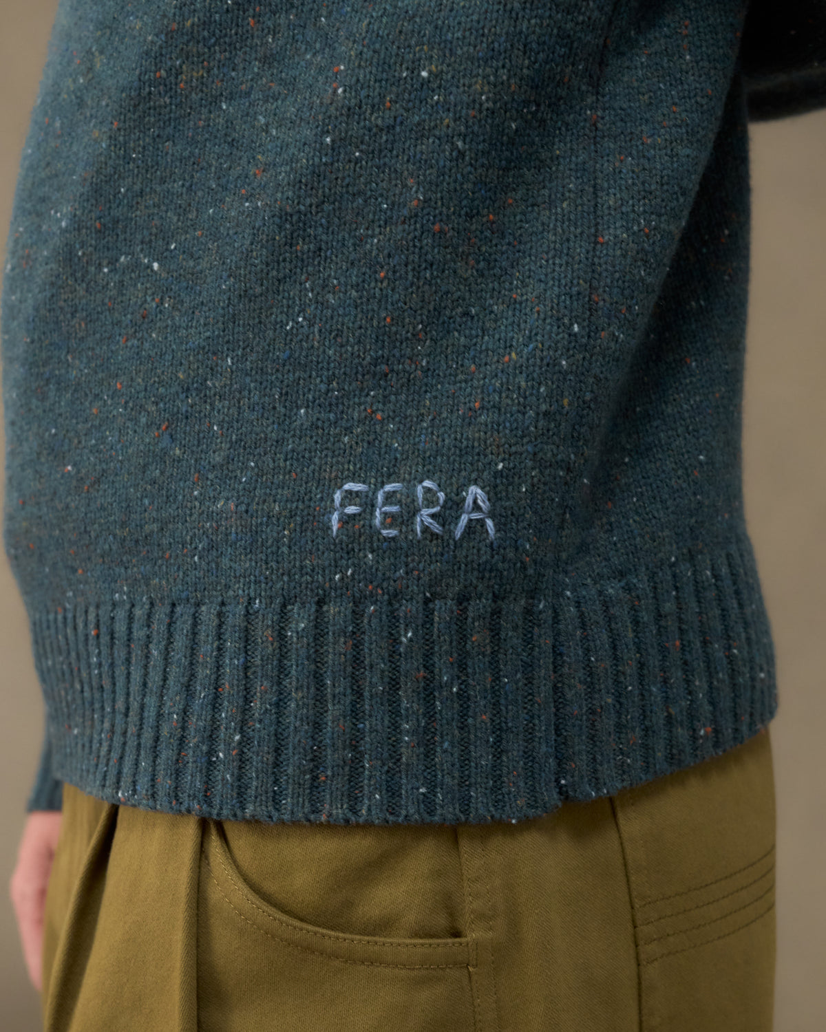 Close-up of the Fera Peewit Knit in Green Donegal showing the hand-embroidered Fera logo and ribbed hem