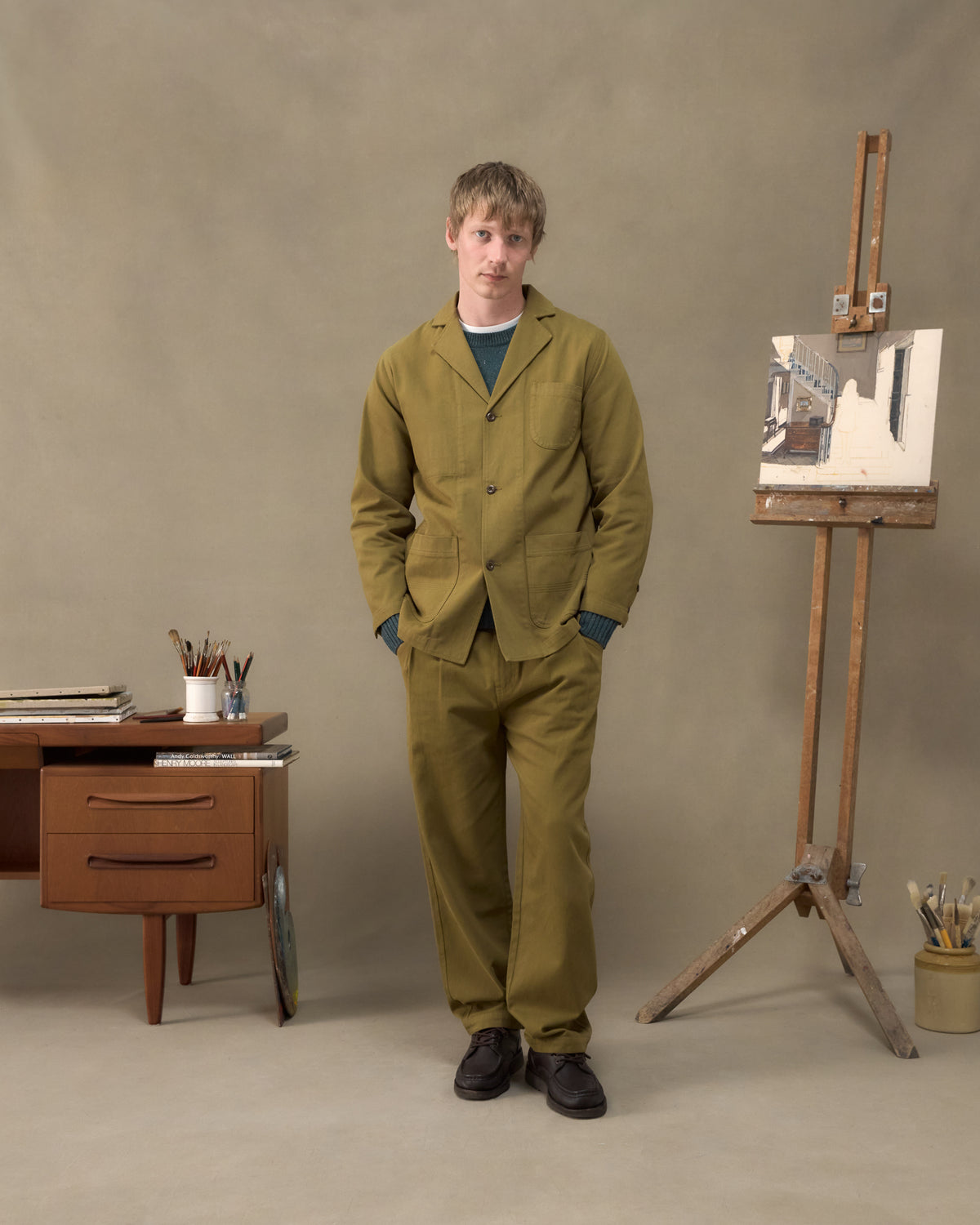 Model wearing the Fera Studio Jacket and Uniform Trousers in Moss, front view beside a painter's easel