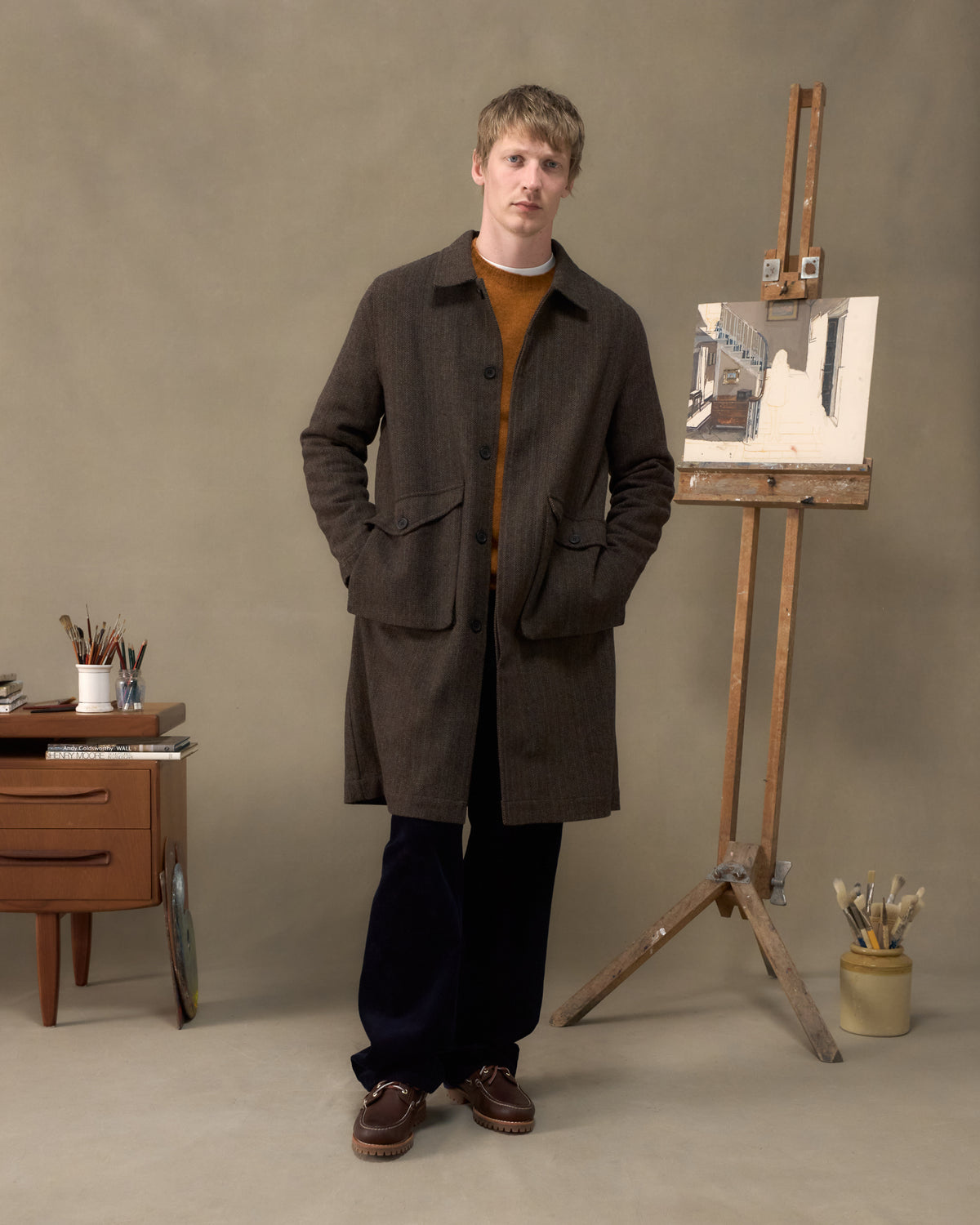 Model wearing the Fera Marwood Coat in Tweed Herringbone over an orange knit in an artist's studio