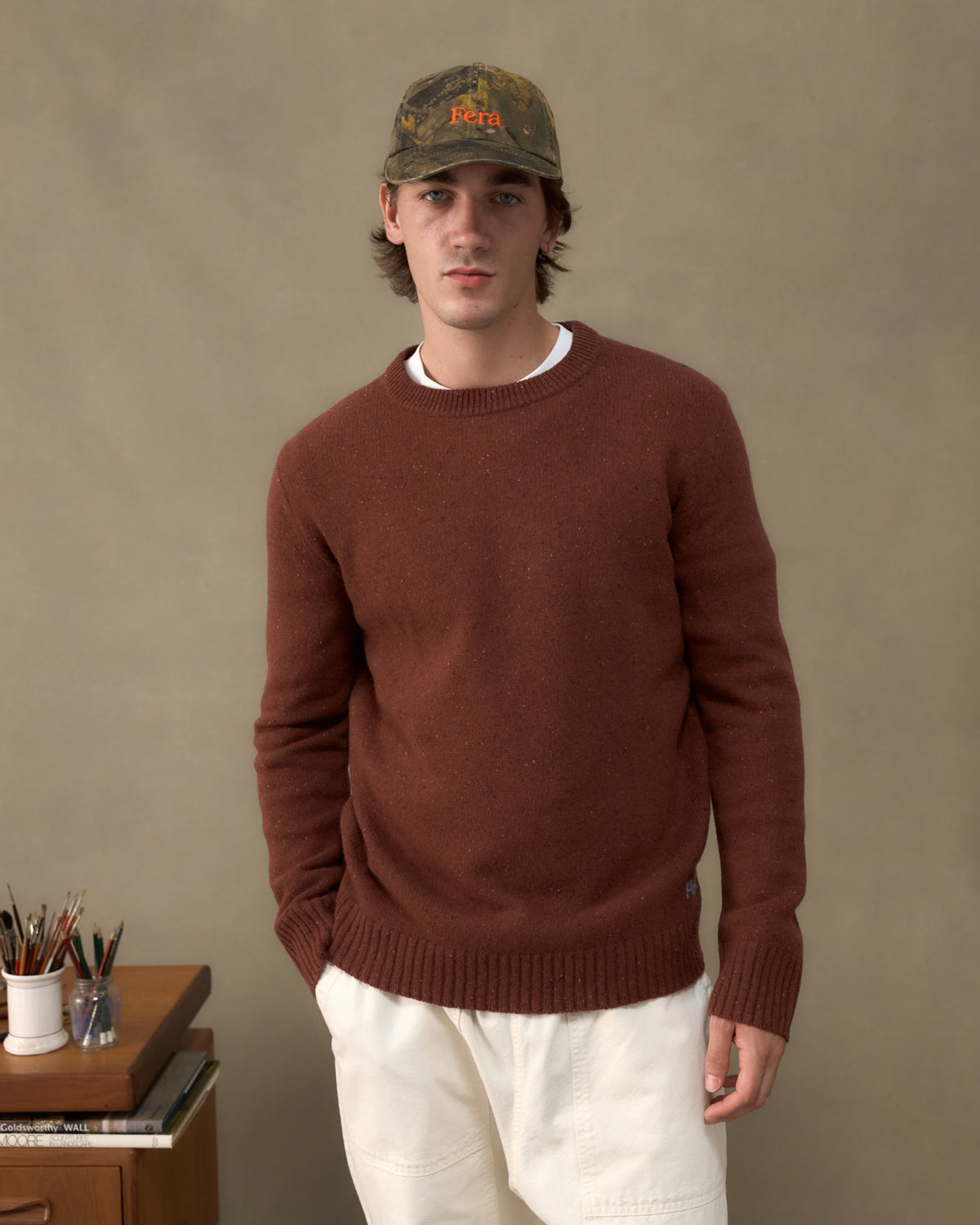 Model wearing the Fera Peewit Knit in Copper with a camouflage Fera cap in an artist's studio