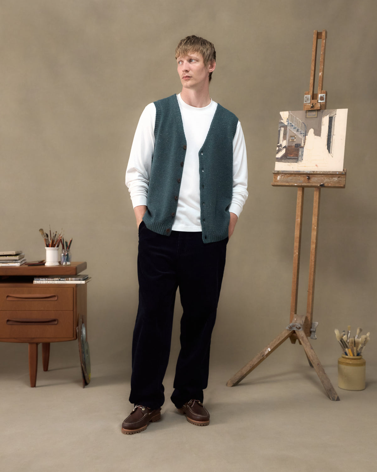 Model wearing the Fera Curlew Merino Vest in Green Donegal with cord trousers in an artist's studio