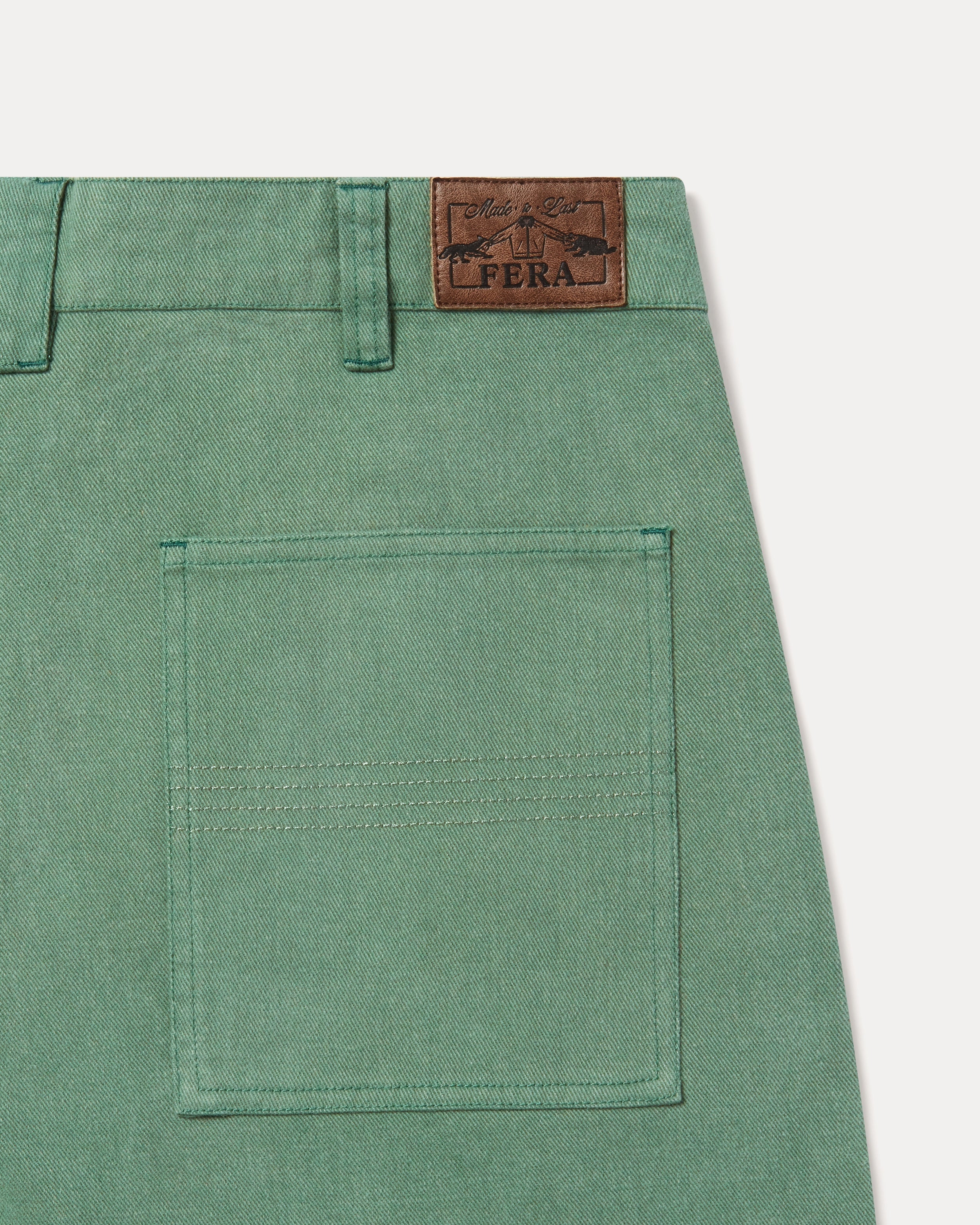 Close-up of the back waistband of green trousers, showing a brown leather patch with the brand name 'FERA' and a rear pocket.